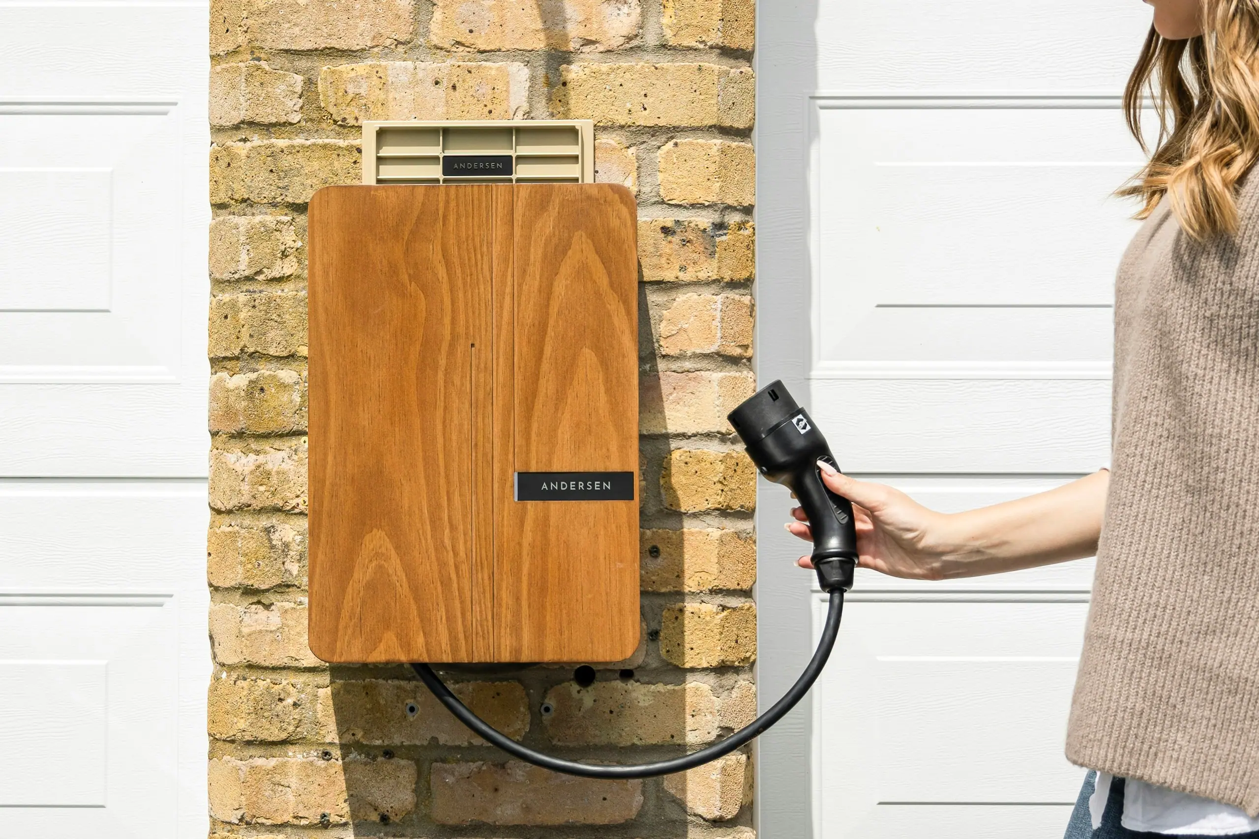 Homeowner lifting the cable from a timber-fronted EV charger on a brick wall